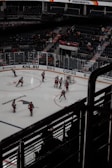 A professional ice hockey game is taking place in an indoor arena. Players in red uniforms are actively participating on the ice rink, with a referee nearby. The stands have a sparse audience, and advertisements are visible along the perimeter.