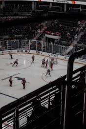 A professional ice hockey game is taking place in an indoor arena. Players in red uniforms are actively participating on the ice rink, with a referee nearby. The stands have a sparse audience, and advertisements are visible along the perimeter.
