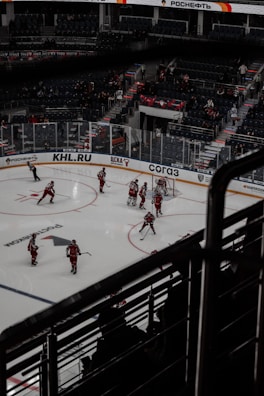 A professional ice hockey game is taking place in an indoor arena. Players in red uniforms are actively participating on the ice rink, with a referee nearby. The stands have a sparse audience, and advertisements are visible along the perimeter.