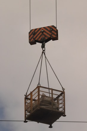 A large industrial hook attached to a crane is suspending a metal cage in the air. The cage appears to be carrying materials or equipment and is held with sturdy chains. The background is a cloudy sky.