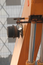 A close-up view of a construction vehicle's hydraulic arm and winch. The metallic cylindrical rods and the orange paint of the machinery stand out prominently. A headlight and cable spool are also visible, with a building in the background showing a pattern of rectangles and diamonds.