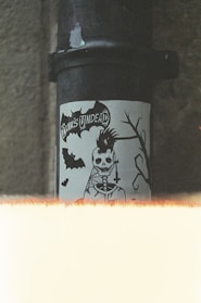 A sticker featuring a skeletal figure with a punk hairstyle and a cross necklace is displayed on a pipe. The text 'Punk's Undead' is visible above the figure. There are bats and a silhouette of a branch in the design. The image has a grainy, vintage look with a light leak effect at the bottom.