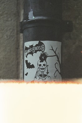 A sticker featuring a skeletal figure with a punk hairstyle and a cross necklace is displayed on a pipe. The text 'Punk's Undead' is visible above the figure. There are bats and a silhouette of a branch in the design. The image has a grainy, vintage look with a light leak effect at the bottom.
