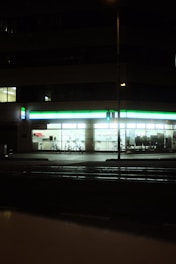 A well-lit convenience store at night with a green and white illuminated sign. The windows reveal shelves stocked with products, and two bicycles are parked outside. A traffic signal glows green on the street corner in the foreground.