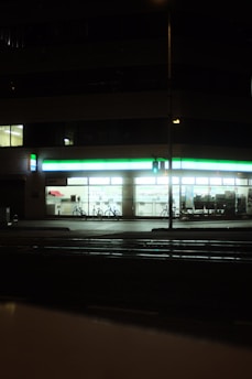 A well-lit convenience store at night with a green and white illuminated sign. The windows reveal shelves stocked with products, and two bicycles are parked outside. A traffic signal glows green on the street corner in the foreground.