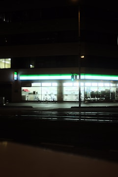A well-lit convenience store at night with a green and white illuminated sign. The windows reveal shelves stocked with products, and two bicycles are parked outside. A traffic signal glows green on the street corner in the foreground.