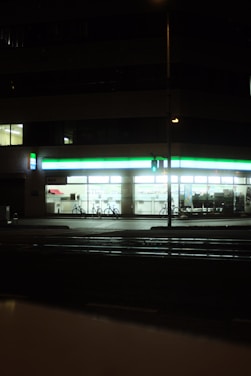 A well-lit convenience store at night with a green and white illuminated sign. The windows reveal shelves stocked with products, and two bicycles are parked outside. A traffic signal glows green on the street corner in the foreground.