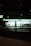 A well-lit convenience store at night with a green and white illuminated sign. The windows reveal shelves stocked with products, and two bicycles are parked outside. A traffic signal glows green on the street corner in the foreground.