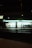 A well-lit convenience store at night with a green and white illuminated sign. The windows reveal shelves stocked with products, and two bicycles are parked outside. A traffic signal glows green on the street corner in the foreground.