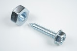 A close-up view of a shiny metallic nut and a screw placed on a plain white surface. The objects are made of steel with a reflective silver finish, emphasizing their industrial design.