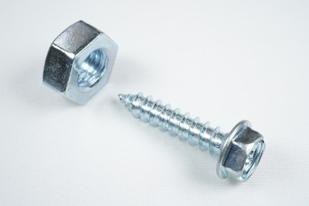 A close-up view of a shiny metallic nut and a screw placed on a plain white surface. The objects are made of steel with a reflective silver finish, emphasizing their industrial design.