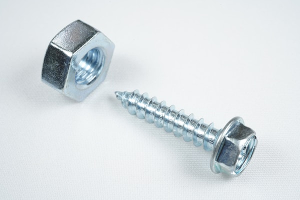 A close-up view of a shiny metallic nut and a screw placed on a plain white surface. The objects are made of steel with a reflective silver finish, emphasizing their industrial design.