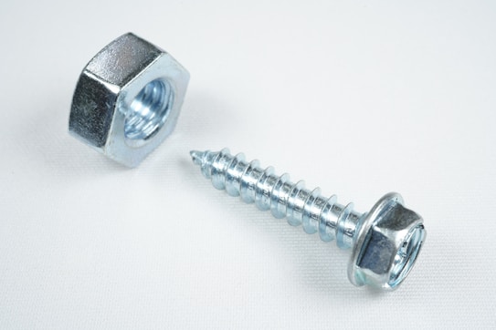 A close-up view of a shiny metallic nut and a screw placed on a plain white surface. The objects are made of steel with a reflective silver finish, emphasizing their industrial design.