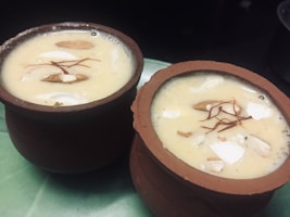 Two clay cups filled with a creamy, beige-colored dessert garnished with slivers of almonds, coconut pieces, and saffron strands. The cups are placed on a green surface, creating a rustic and traditional presentation.