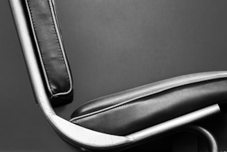 A close-up of a modern chair featuring black leather upholstery with visible stitches and a metallic frame. The image highlights the sleek lines and minimalist design of the chair against a plain background.