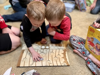 Two young children are engrossed in playing a board game on the floor, surrounded by family members. They are leaning over the game board with focused expressions. One child is wearing a red shirt and the other is wearing black. In the background, there are people seated on the floor and various wrapped and unwrapped gifts, indicating a festive occasion.
