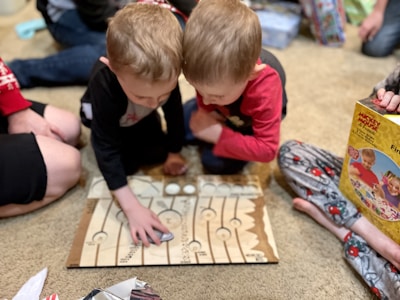 Two young children are engrossed in playing a board game on the floor, surrounded by family members. They are leaning over the game board with focused expressions. One child is wearing a red shirt and the other is wearing black. In the background, there are people seated on the floor and various wrapped and unwrapped gifts, indicating a festive occasion.