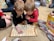 Two young children are engrossed in playing a board game on the floor, surrounded by family members. They are leaning over the game board with focused expressions. One child is wearing a red shirt and the other is wearing black. In the background, there are people seated on the floor and various wrapped and unwrapped gifts, indicating a festive occasion.