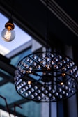 A decorative light fixture with a vintage-style bulb hanging from a ceiling. The bulb is lit, showcasing a clear filament design, and is surrounded by an ornate chandelier structure with numerous small lights. The background consists of a glass ceiling with visible structural elements, creating a contemporary indoor ambiance.