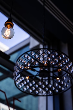 A decorative light fixture with a vintage-style bulb hanging from a ceiling. The bulb is lit, showcasing a clear filament design, and is surrounded by an ornate chandelier structure with numerous small lights. The background consists of a glass ceiling with visible structural elements, creating a contemporary indoor ambiance.