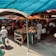 A bustling outdoor market with various stalls selling fresh produce, including tomatoes, cucumbers, and other vegetables. Several people are shopping and interacting with vendors under colorful tarps providing shade. Brightly colored canopies in blue, orange, and white add vibrancy to the atmosphere. Some shoppers carry bags of purchased goods as they navigate the busy marketplace.
