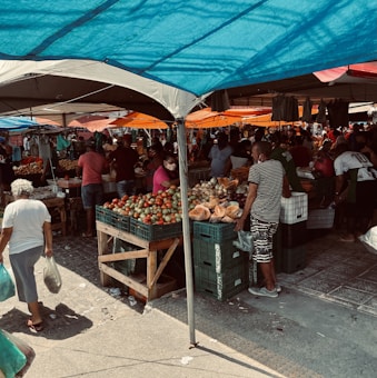 A bustling outdoor market with various stalls selling fresh produce, including tomatoes, cucumbers, and other vegetables. Several people are shopping and interacting with vendors under colorful tarps providing shade. Brightly colored canopies in blue, orange, and white add vibrancy to the atmosphere. Some shoppers carry bags of purchased goods as they navigate the busy marketplace.