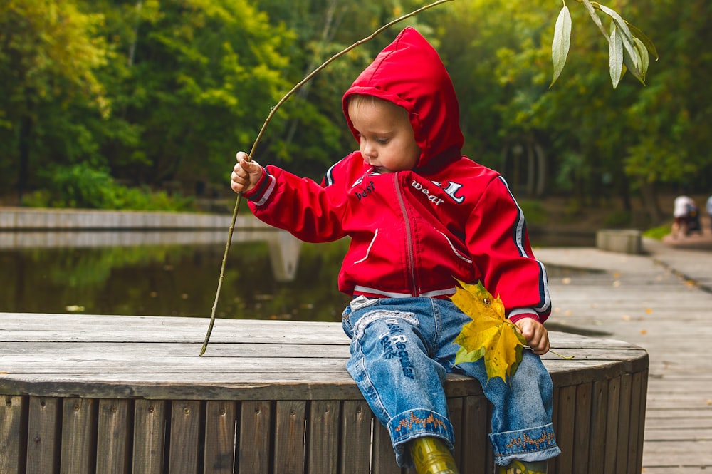boy in red hoodie and blue denim jeans sitting on wooden dock during daytime