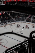 Aerial view of a professional ice hockey game in progress, with players in red and white jerseys gathered at the center faceoff circle. The arena is partially filled with spectators and features large digital displays and advertisements around the perimeter of the rink.