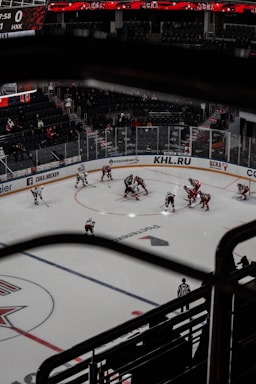 Aerial view of a professional ice hockey game in progress, with players in red and white jerseys gathered at the center faceoff circle. The arena is partially filled with spectators and features large digital displays and advertisements around the perimeter of the rink.