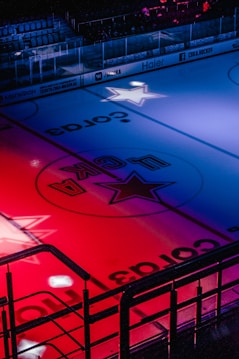 An ice hockey rink is illuminated with vibrant red and blue lights, casting colorful reflections on the ice. The rink features advertising logos and a central star symbol. Empty stands are visible in the background, and the atmosphere suggests anticipation.