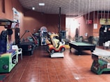 An indoor arcade area featuring various gaming machines, including an air hockey table and a ride-on toy car. The room has a tiled floor and several colorful gaming devices scattered around. Chairs and arcade signage are visible, creating a playful and entertainment-focused atmosphere.