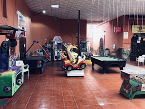 An indoor arcade area featuring various gaming machines, including an air hockey table and a ride-on toy car. The room has a tiled floor and several colorful gaming devices scattered around. Chairs and arcade signage are visible, creating a playful and entertainment-focused atmosphere.