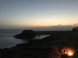 The image captures a coastal scene at dusk, with a large rock formation jutting out into a calm sea. The sky is a gradient of soft colors, transitioning from a pale orange near the horizon to a deeper blue further up. A crescent moon is faintly visible. In the foreground, a group of people are gathered around a bright, warm fire.