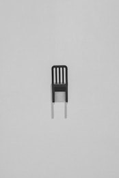 A single, minimalist black chair with a slatted backrest set against a light gray, textured background. The chair casts a subtle shadow beneath it, adding depth to the stark composition.