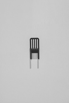 A single, minimalist black chair with a slatted backrest set against a light gray, textured background. The chair casts a subtle shadow beneath it, adding depth to the stark composition.