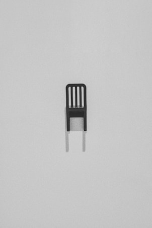 A single, minimalist black chair with a slatted backrest set against a light gray, textured background. The chair casts a subtle shadow beneath it, adding depth to the stark composition.
