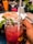 A close-up of a vibrant pink beverage in a mason jar garnished with mint leaves, held by a hand with a striped straw. In the background, there is a plate of food and a glass with a yellow-orange drink. Other elements include a water glass, some greenery, and subtle reflections indicating an indoor setting.