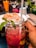 A close-up of a vibrant pink beverage in a mason jar garnished with mint leaves, held by a hand with a striped straw. In the background, there is a plate of food and a glass with a yellow-orange drink. Other elements include a water glass, some greenery, and subtle reflections indicating an indoor setting.