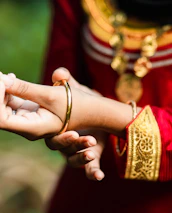 A close-up of a person wearing traditional attire featuring intricate gold embroidery and a red fabric. The individual is adjusting a gold bangle on their wrist. The outfit is complemented by ornate jewelry, including a pendant necklace.