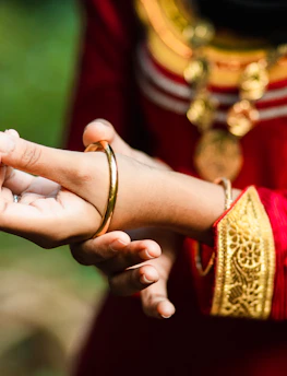 A close-up of a person wearing traditional attire featuring intricate gold embroidery and a red fabric. The individual is adjusting a gold bangle on their wrist. The outfit is complemented by ornate jewelry, including a pendant necklace.