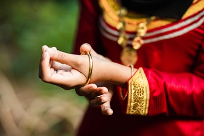 A close-up of a person wearing traditional attire featuring intricate gold embroidery and a red fabric. The individual is adjusting a gold bangle on their wrist. The outfit is complemented by ornate jewelry, including a pendant necklace.