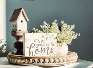 A decorative still life featuring a small wooden birdhouse with a rustic design, placed next to a sign that reads 'it's so good to be home' with a cursive style. Beside it is a vase filled with delicate white flowers, all sitting on an ornate, round wooden tray with carved detailing. Soft lighting casts gentle shadows, creating a calm and cozy atmosphere.