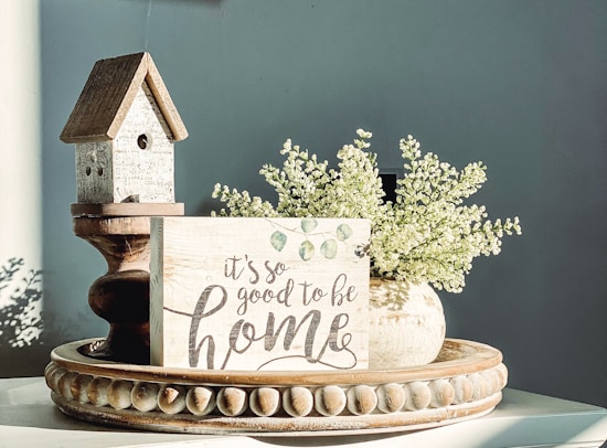A decorative still life featuring a small wooden birdhouse with a rustic design, placed next to a sign that reads 'it's so good to be home' with a cursive style. Beside it is a vase filled with delicate white flowers, all sitting on an ornate, round wooden tray with carved detailing. Soft lighting casts gentle shadows, creating a calm and cozy atmosphere.