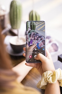 A person is holding a smartphone displaying several photo cards of people. The phone screen shows a camera interface with various options. In the background, there is a blurred image of a cactus and a coffee cup on the table. The person is wearing a yellow and white checkered scrunchie.