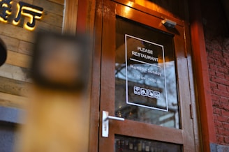 A wooden framed glass door with signage indicating the name 'Please Restaurant'. The operating hours are Monday to Sunday, from 11:30 am to midnight. The door has icons suggesting free Wi-Fi, wheelchair accessibility, and other amenities. A brick wall and wooden paneling are nearby.