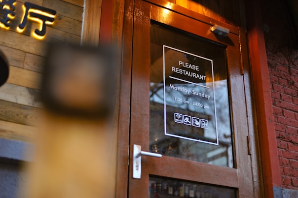 A wooden framed glass door with signage indicating the name 'Please Restaurant'. The operating hours are Monday to Sunday, from 11:30 am to midnight. The door has icons suggesting free Wi-Fi, wheelchair accessibility, and other amenities. A brick wall and wooden paneling are nearby.