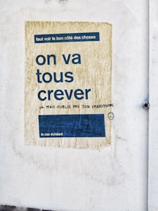 A faded poster on a textured wall with French text in large, bold letters. The background of the poster is a worn, yellowish color with a blue rectangle containing smaller text. Beneath the main message, there is handwritten graffiti in black ink, adding a humorous remark and a smiley face drawing.