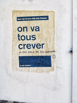 A faded poster on a textured wall with French text in large, bold letters. The background of the poster is a worn, yellowish color with a blue rectangle containing smaller text. Beneath the main message, there is handwritten graffiti in black ink, adding a humorous remark and a smiley face drawing.