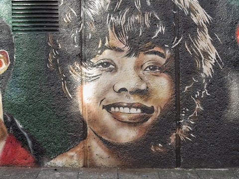 A detailed mural on a textured wall depicts the smiling face of a person with curly hair. The facial expression is joyful and the artwork uses light and shadow effectively to create a realistic effect. The surrounding area is adorned with dark greens and light browns, contributing to the overall composition.