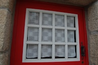 A window set in a red wooden frame, featuring a grid pattern with white lace curtains in each section. The window is bordered by textured stone walls, and an ornate black metal handle is visible on the right side.